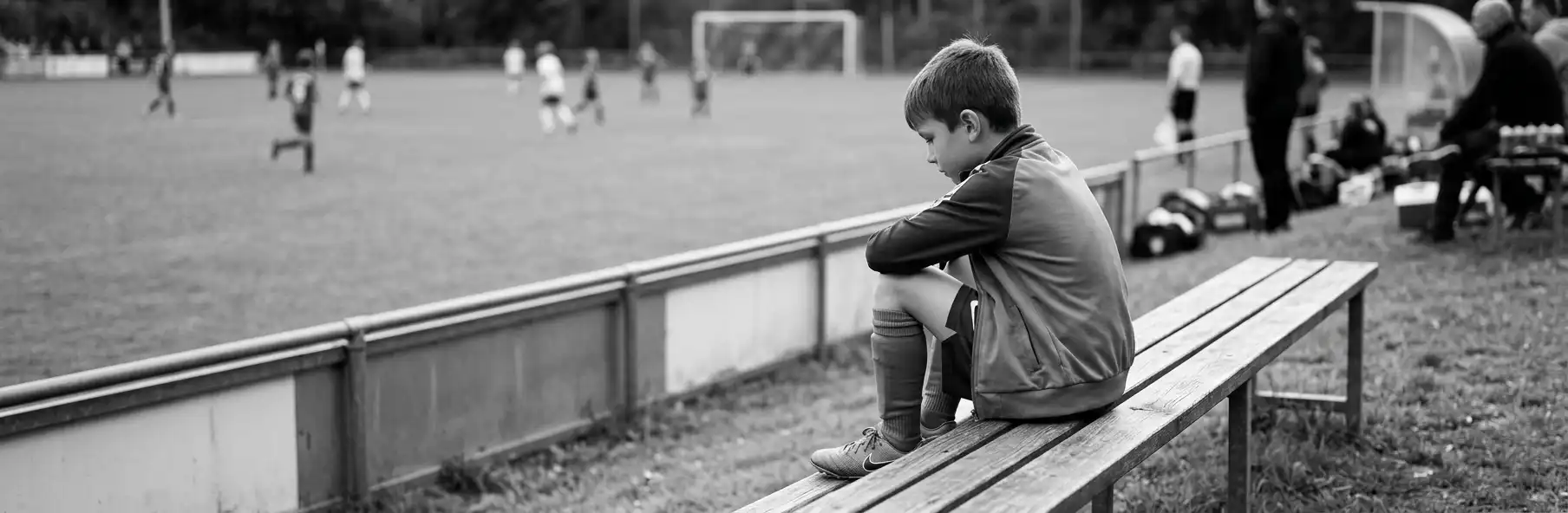 Parent watching a youth football match from the sideline while their child sits on the substitutes' bench
