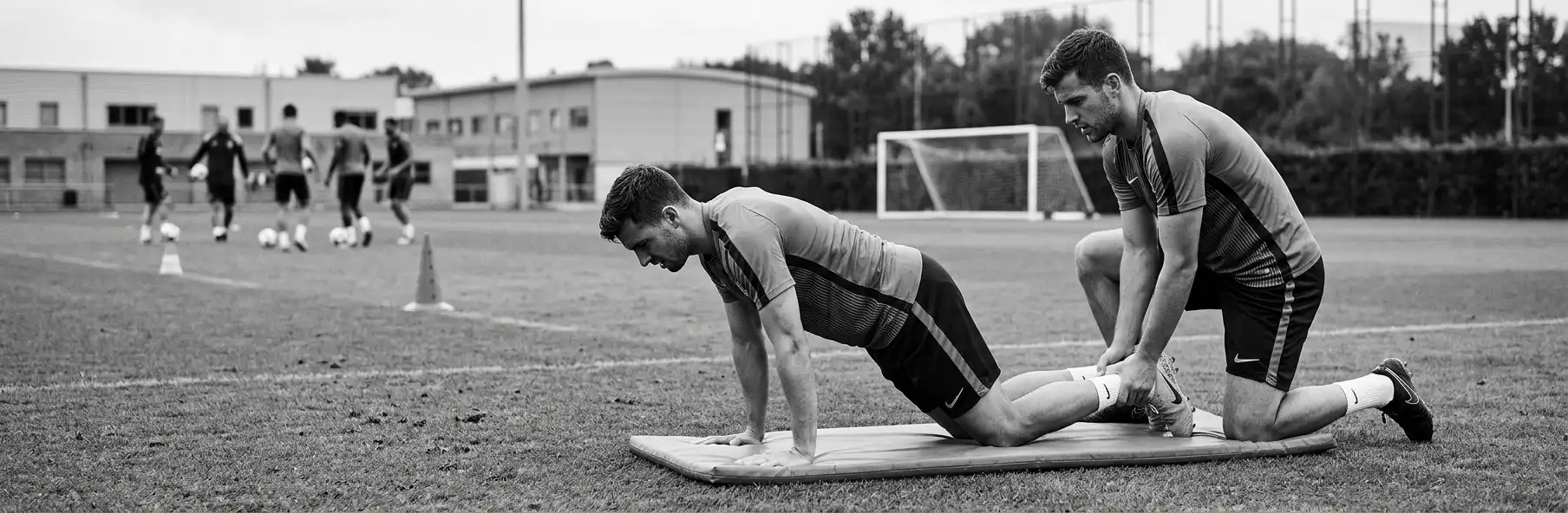 Two football players performing the Nordic hamstring curl on the pitch, one holding the other's ankles